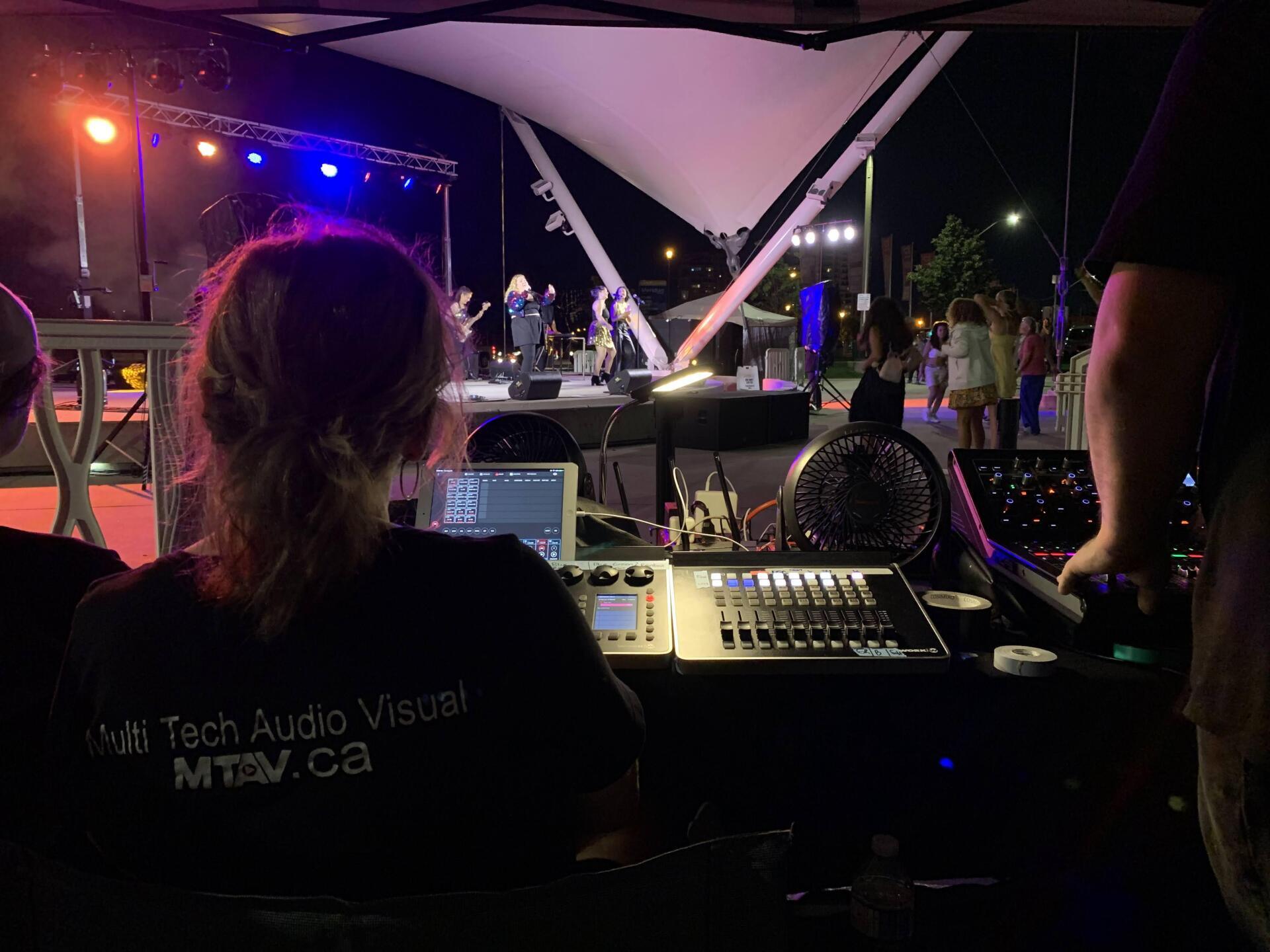 Person at soundboard controls music for a band performing on an outdoor stage at night.