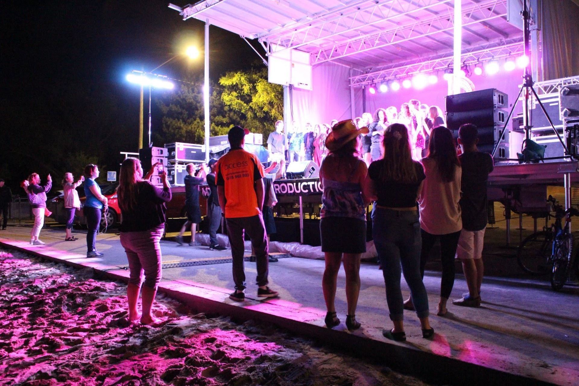 People watching a performance on an outdoor stage at night, lit with pink and purple lights.