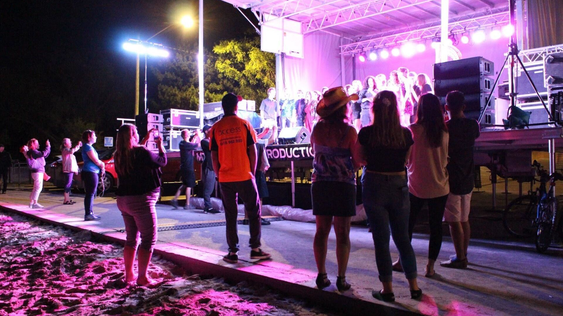 People watch a band perform on an outdoor stage at night. Stage lit with pink lights.