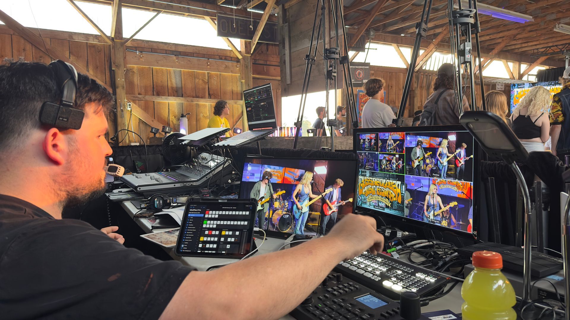 Man operating video equipment inside a barn with band on monitors. Beverage in foreground.