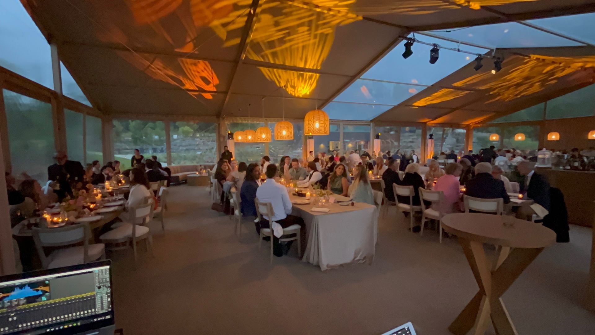 Wedding reception inside a tent, with guests seated at tables. Warm lighting illuminates the scene.