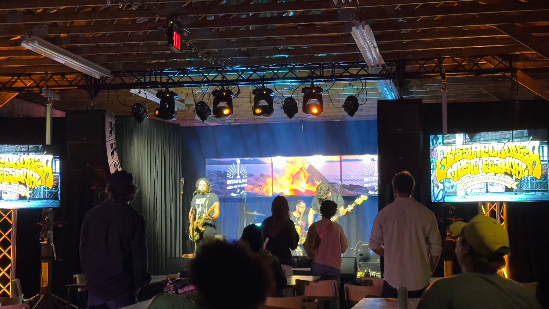 Band performing on stage with screens displaying visuals, audience watching.