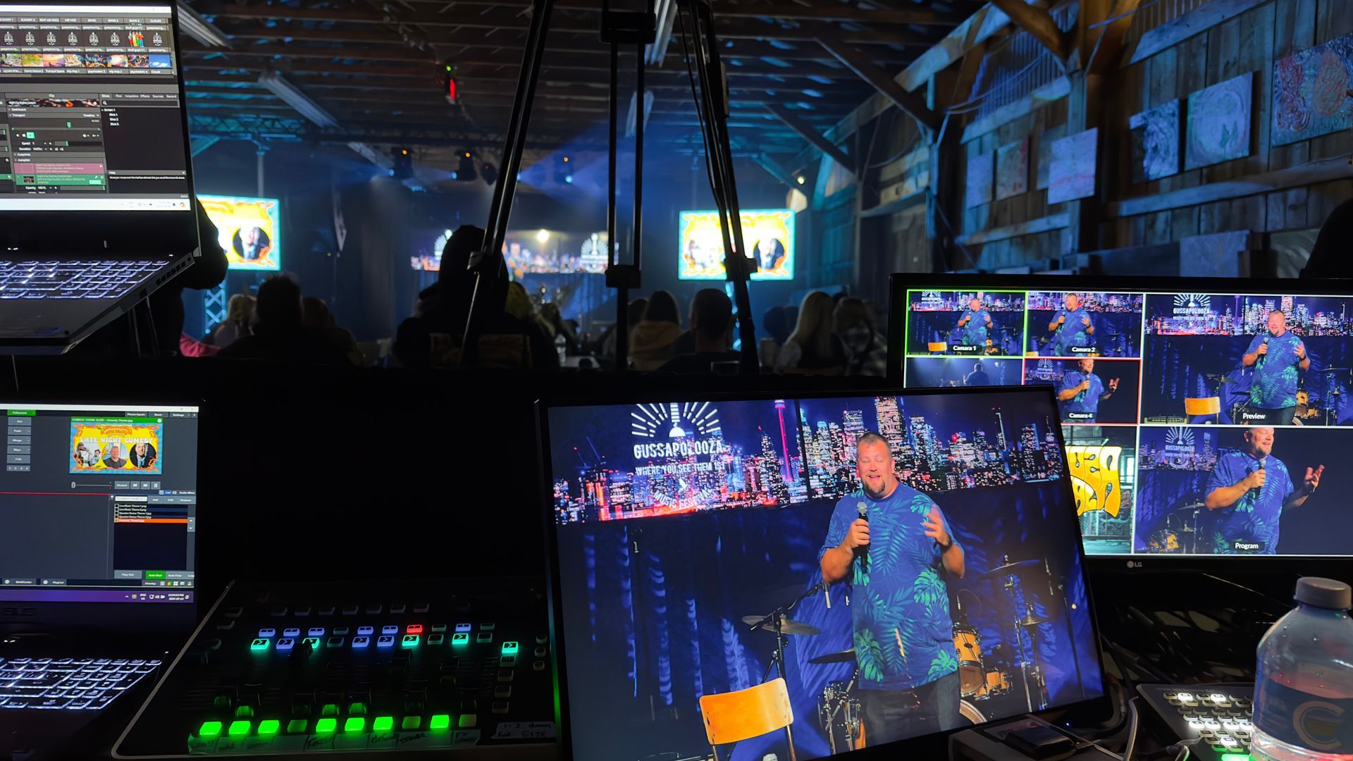 Live event production control room; monitors show stage performance. Dimly lit; consoles and equipment visible.