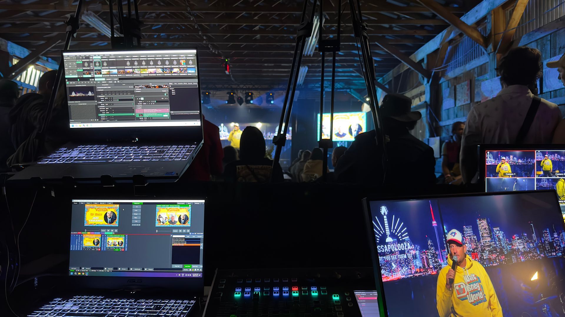Laptops and screens displaying live event footage in a dimly lit venue. Stage and audience visible in the background.