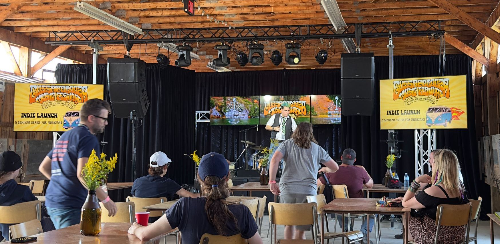 A band plays on stage at an outdoor venue with an audience at tables.