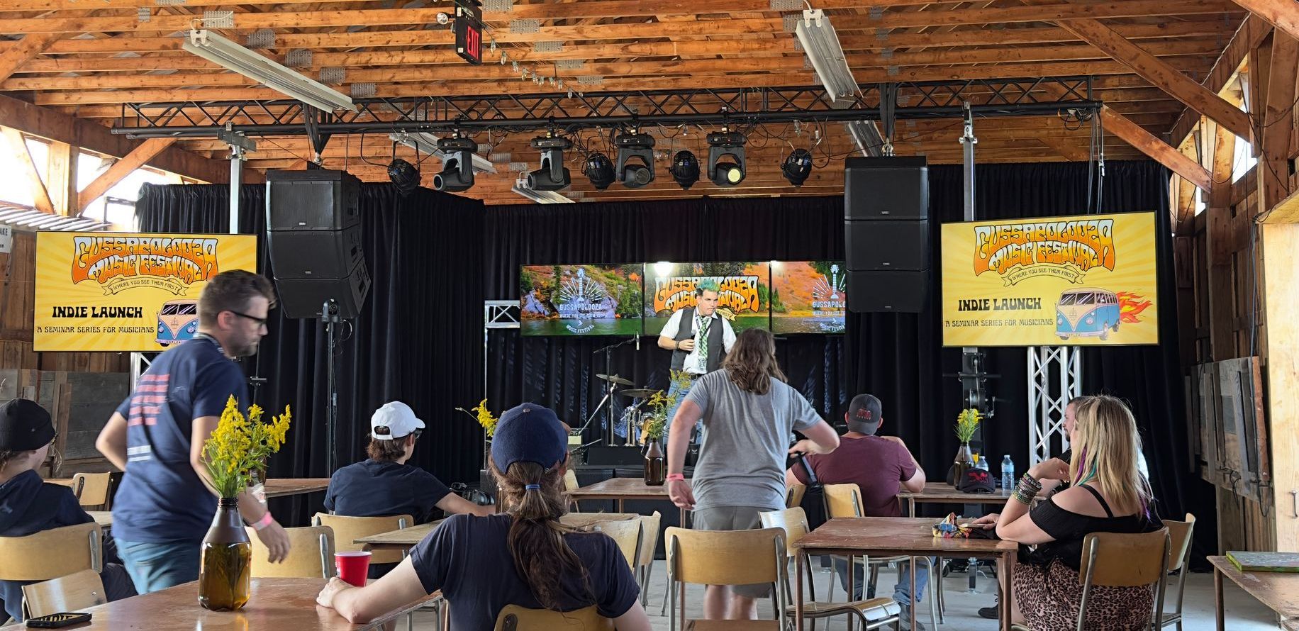 People seated at tables watch a performance on stage in a rustic venue with wooden beams.