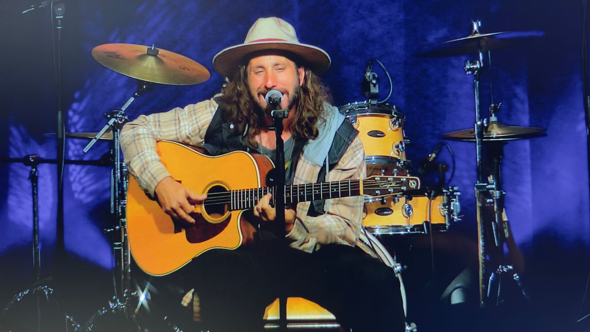 Man with hat sings and plays acoustic guitar on stage, drums in the background.