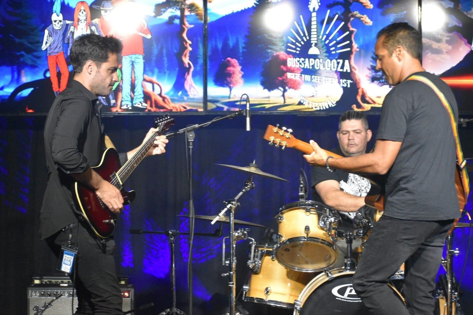 Band performing on stage with guitars and drums. Background features a projection of a landscape.