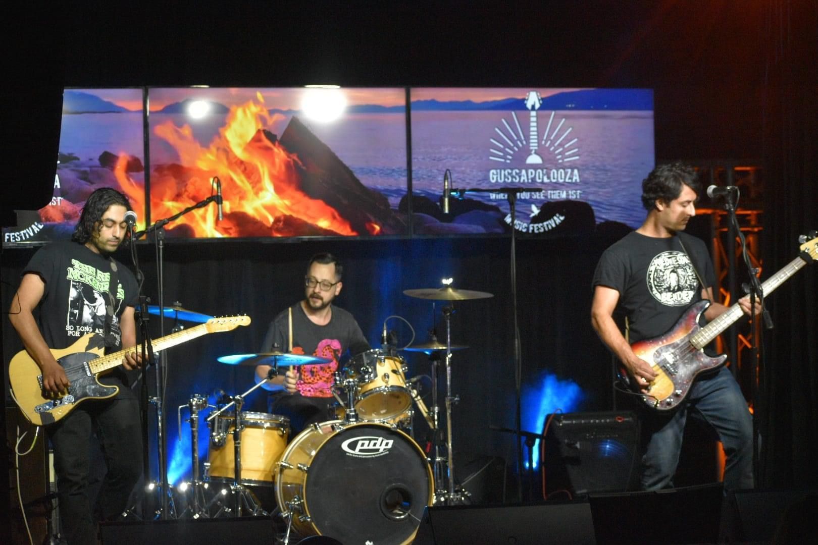 Band performing on stage with a campfire backdrop. Guitarists and drummer playing instruments. Stage lit with blue spotlights.