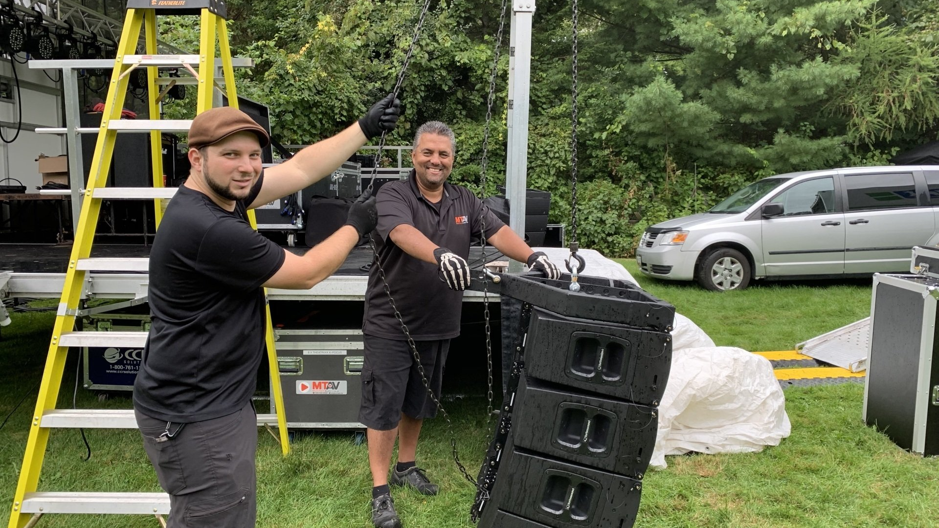 Two men assembling speaker system outdoors near a ladder, van, and equipment cases.