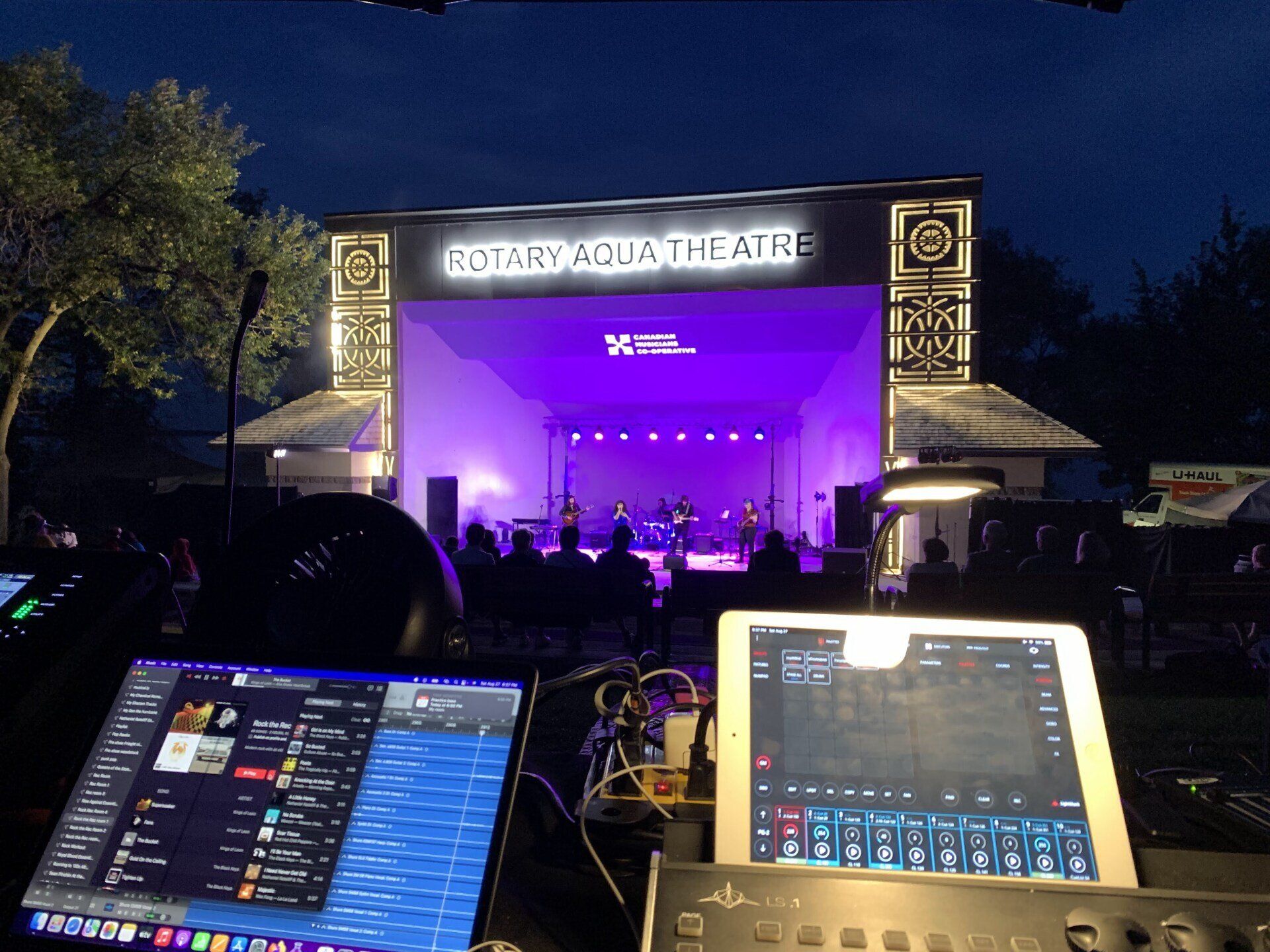 A band performs on stage at the Rotary Aqua Theatre, illuminated in purple light. Mixing consoles in the foreground.