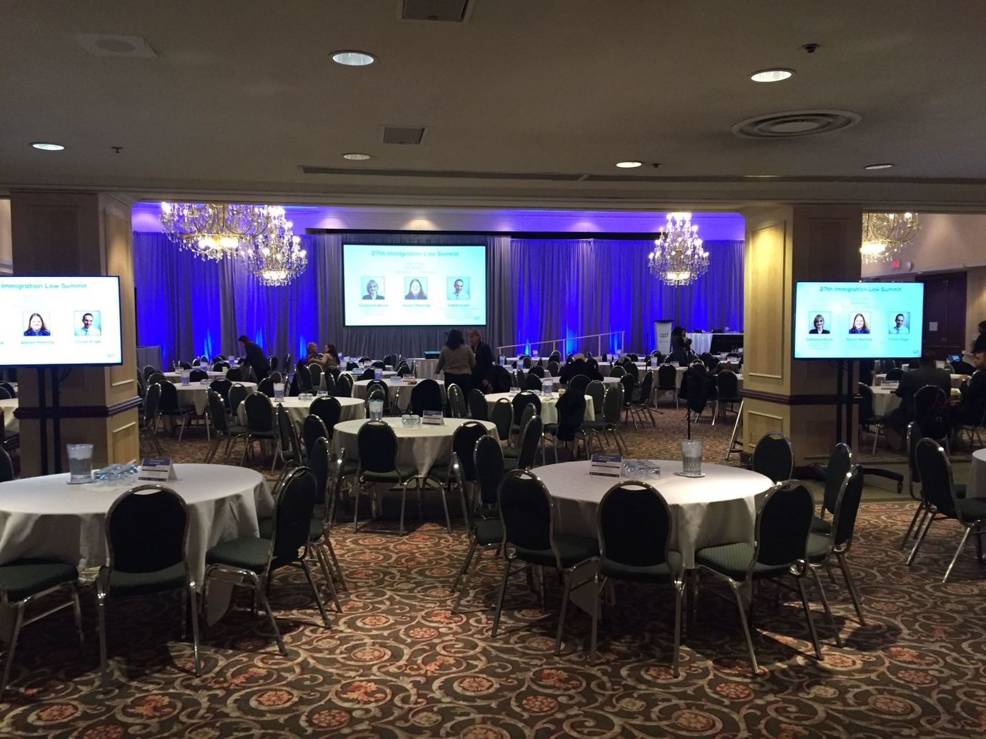 A banquet hall set for a conference with round tables, screens, and a stage with a presentation.
