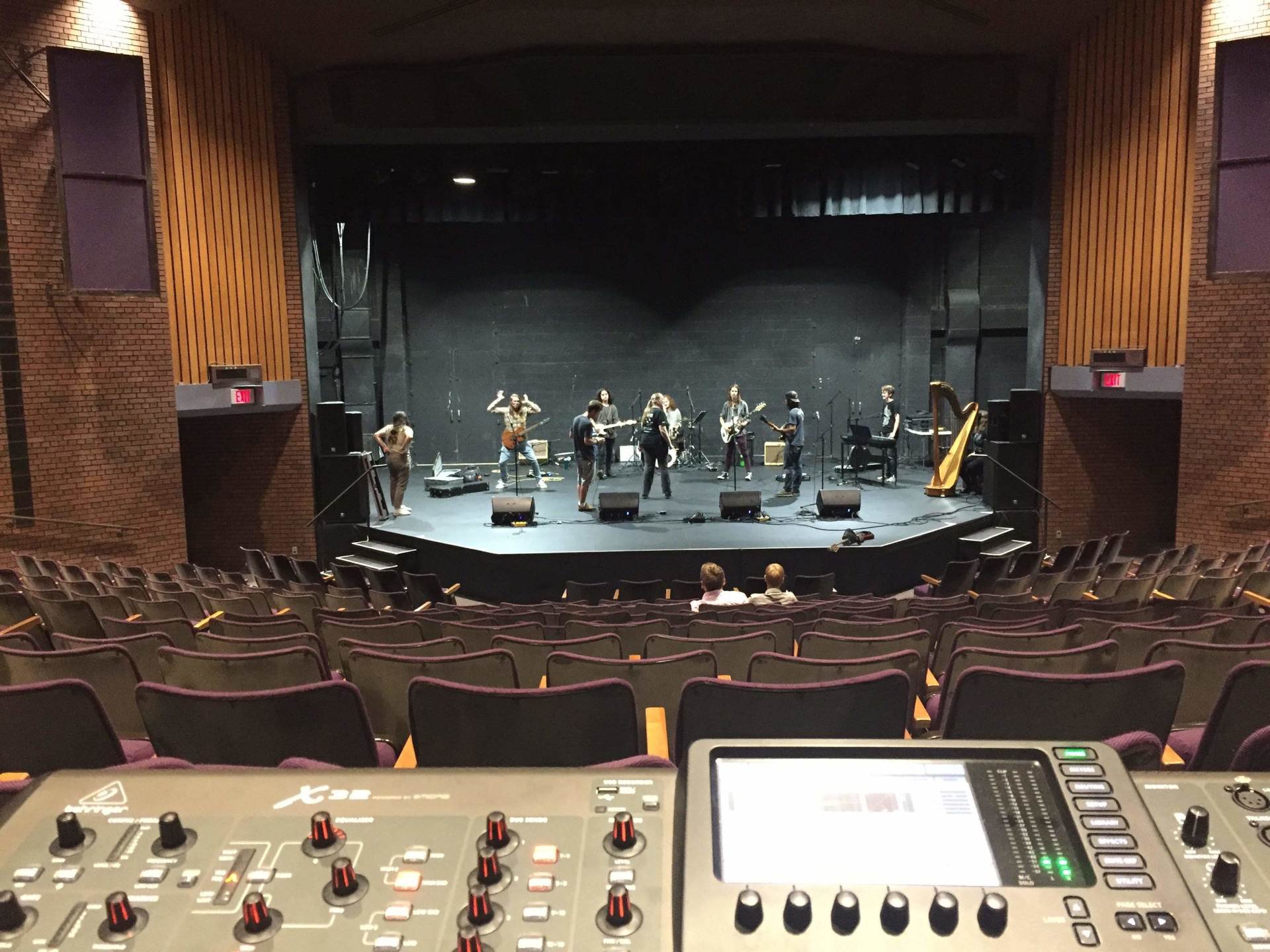 Band on a stage in a theater, seen from the soundboard. The stage has multiple musicians performing.
