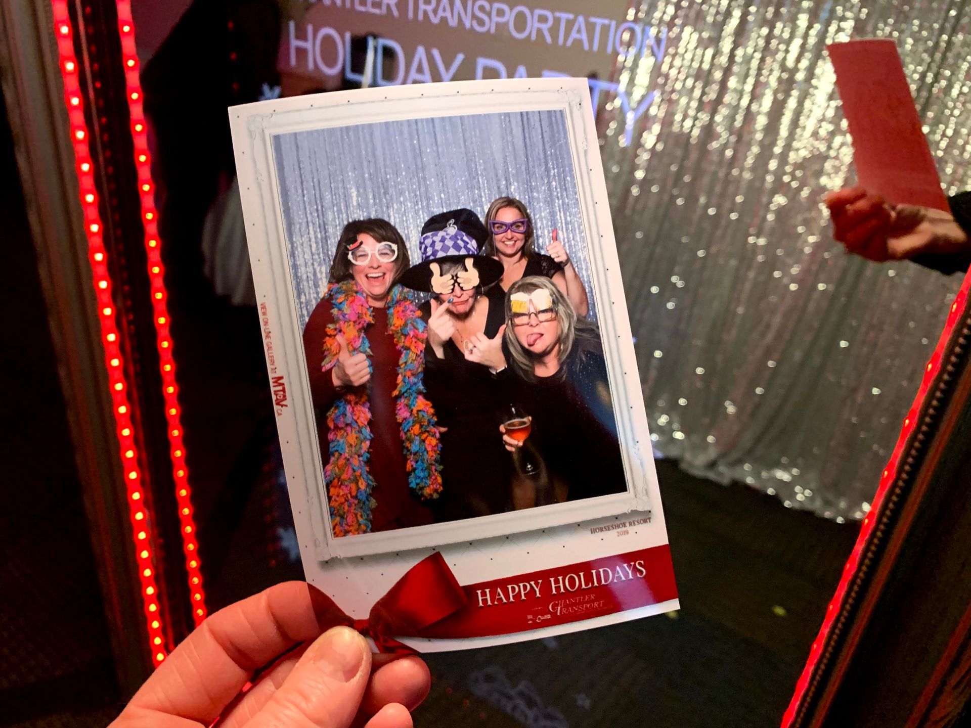 Photo booth picture of five people smiling, holding a “Happy Holidays” print, taken at a party with a sparkly backdrop.