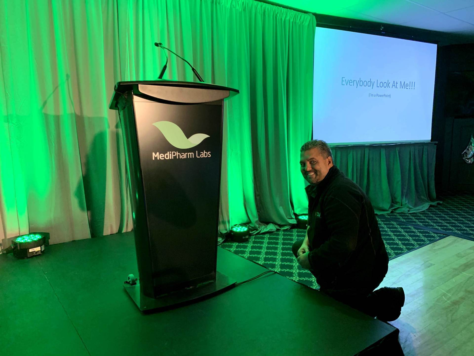 Man kneels beside a black podium with logo. Green lighting illuminates stage and backdrop.