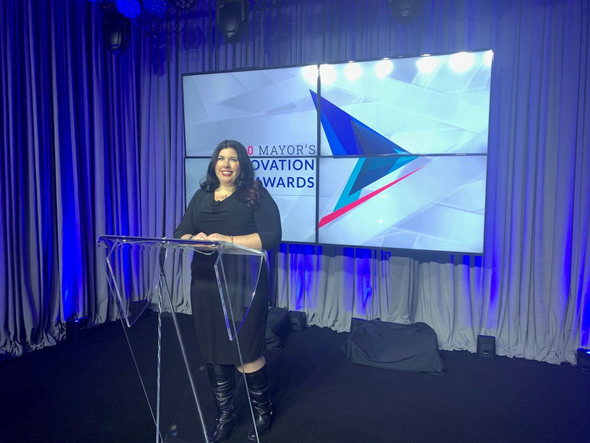 Woman standing at podium, presenting at an awards ceremony. Blue backdrop with screen displaying event logo.