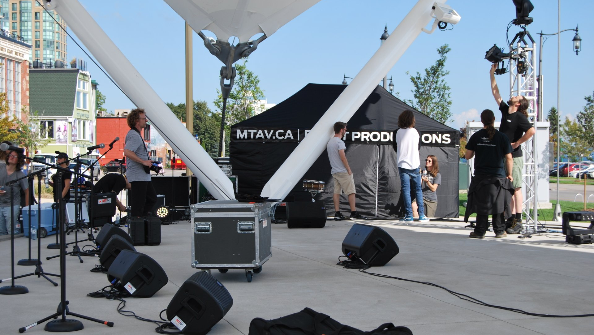 Outdoor stage setup with crew working. Speakers, lights, tent backdrop. Sunny day.