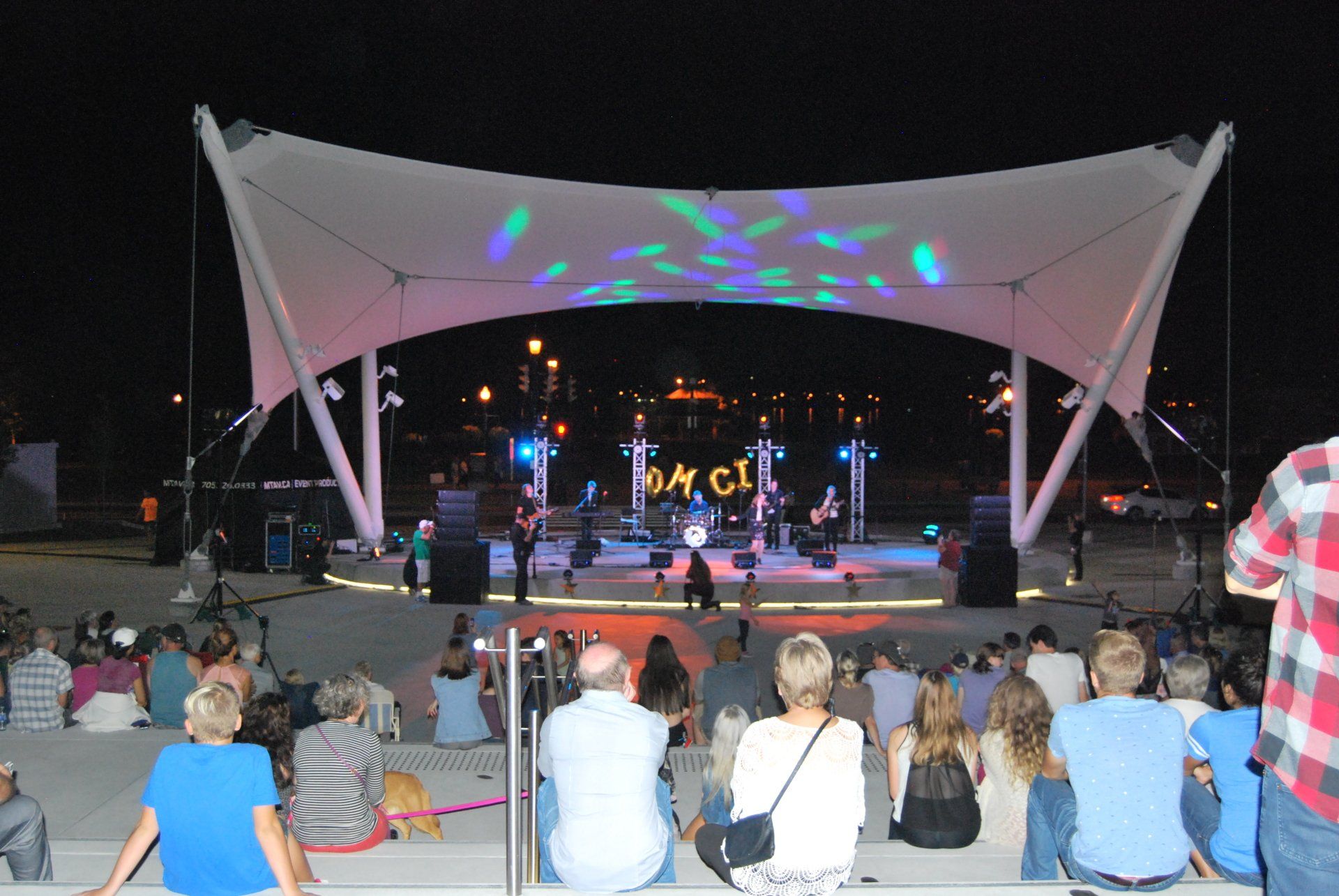 Band performs on an outdoor stage at night, audience watching. Stage lit with colorful lights, white canopy.