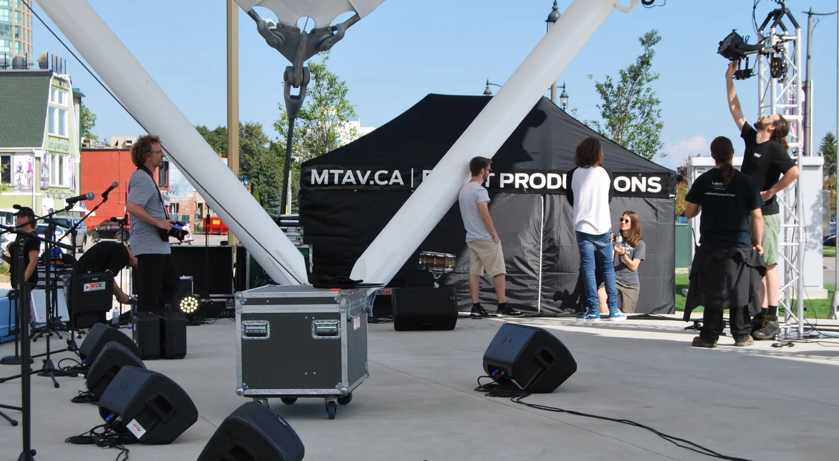 People setting up stage equipment, including speakers and lights, outdoors under a white canopy on a sunny day.
