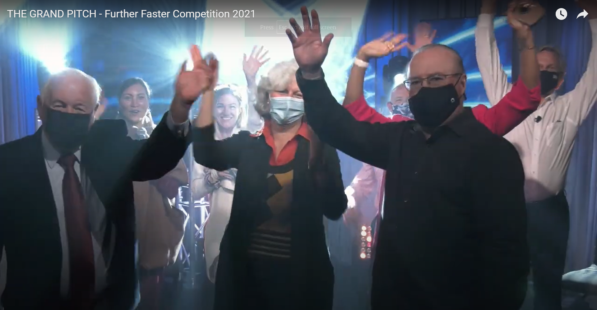 People in masks raising arms in celebration on a stage, blue and white background, spotlights.