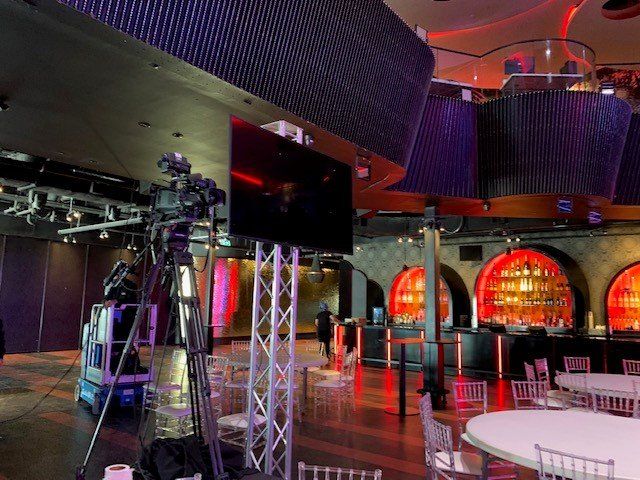 A camera setup with a large screen in a venue with tables, chairs, and a bar, all illuminated by colorful lights.