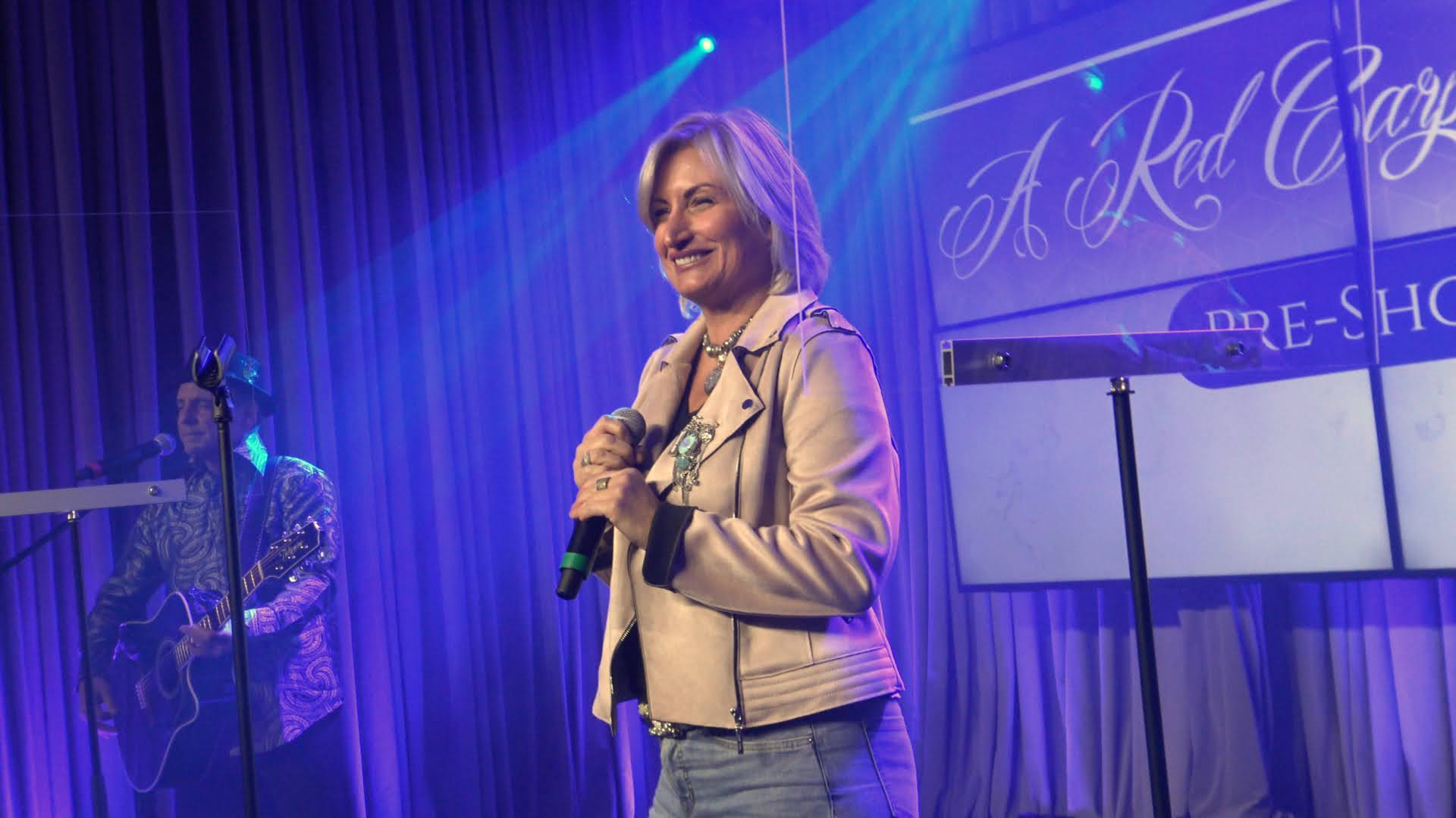 Woman on stage holding a microphone, beige jacket and jeans. Blue stage lighting, background text.