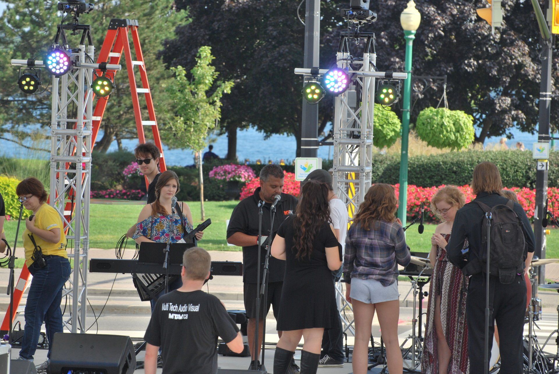 Band preparing to perform on outdoor stage, lake in background, people milling about.