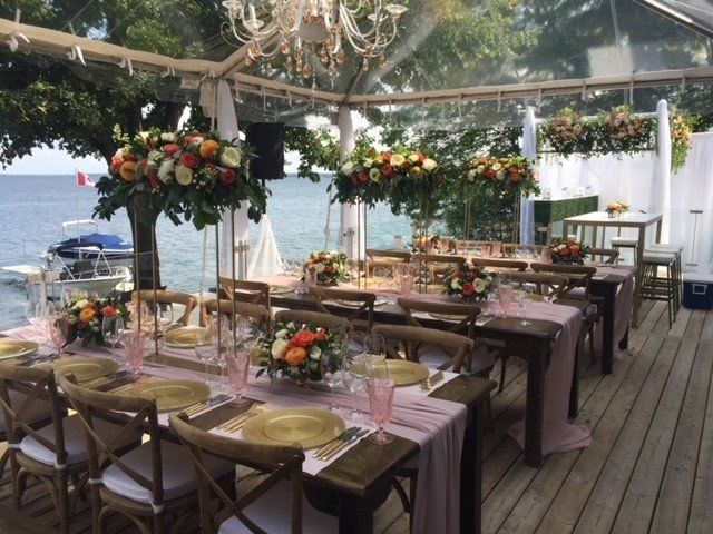 Outdoor dining tables set with floral centerpieces, under a clear tent, overlooking the water.