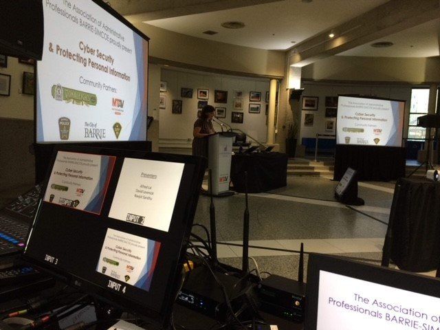 A woman presents on cybersecurity at a conference. Projected slides and monitors display information.