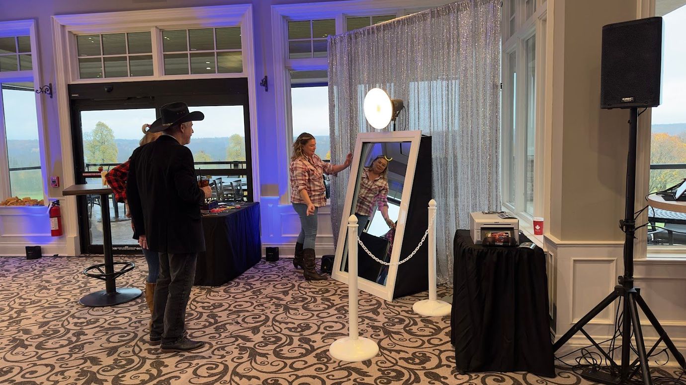 lady standing infront of a photobooth that looks like a large mirror. man in hat standing infront. white curtains in the back and white stanchions to the side. A black skirted table has the PA mixing board on it.