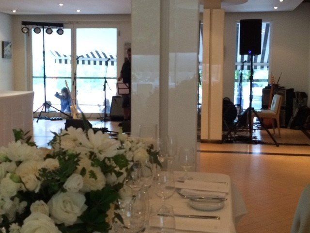 Restaurant interior with white flowers, tables set for a meal, and a band setting up with equipment near a window.
