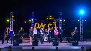 Band performing on outdoor stage with blue lighting and logo backdrop.