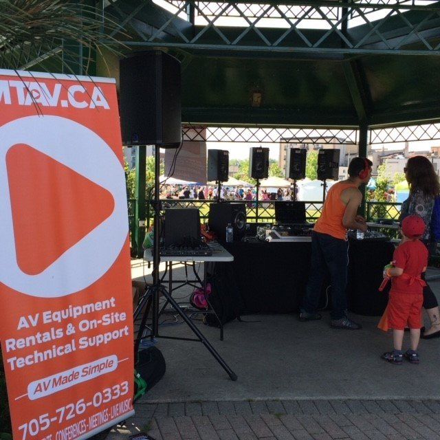 An AV equipment rental booth at an outdoor event. Sign with orange logo. People interact near the speakers.
