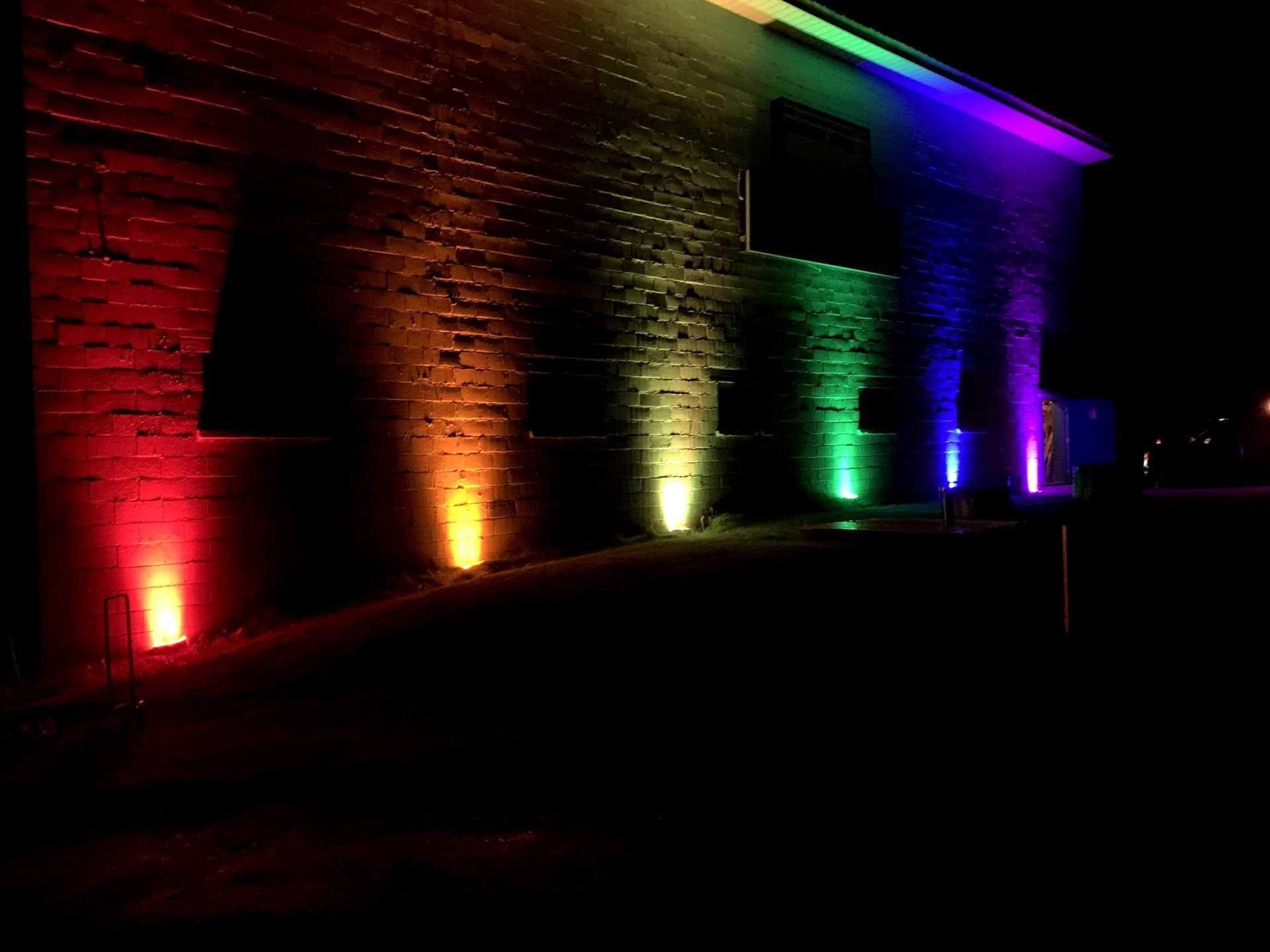 A brick building illuminated at night with colorful uplights: red, orange, yellow, green, blue, and purple.