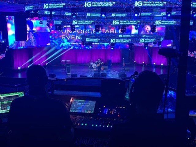 Two people at a soundboard, stage lit with purple and white lights, backdrop of 