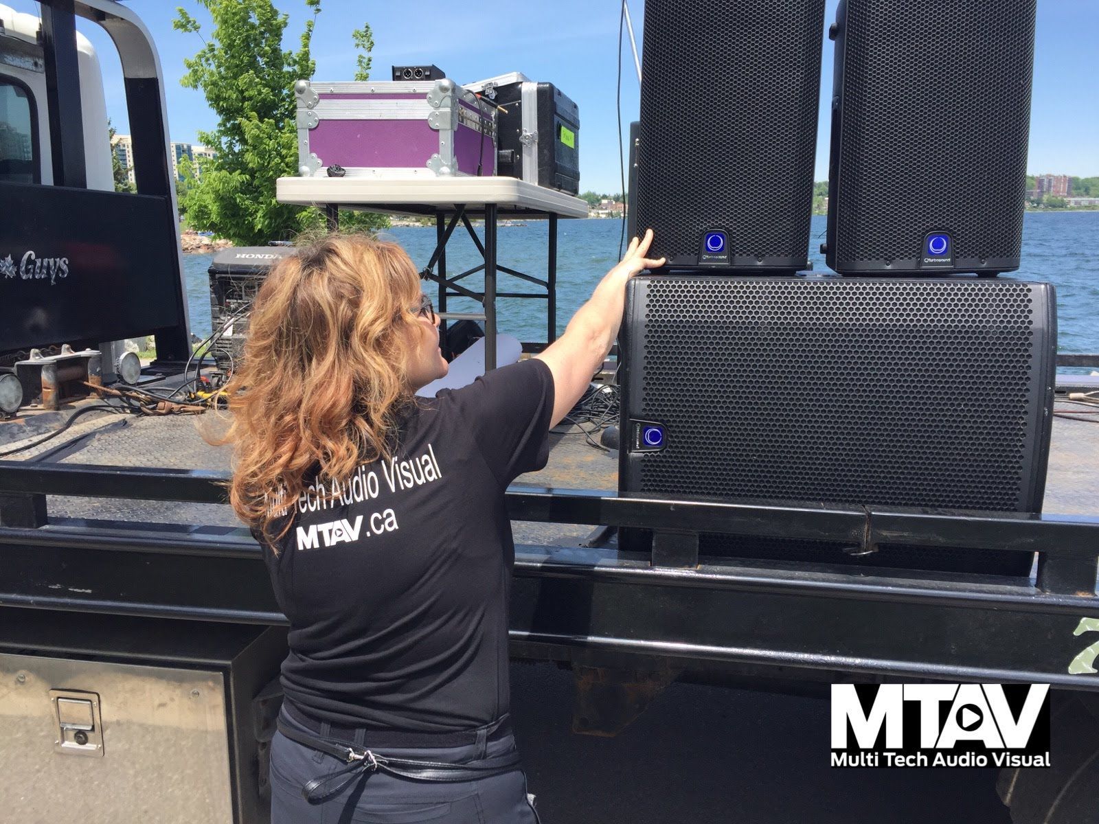 Woman arranging black speakers on a truck bed with a waterfront background.