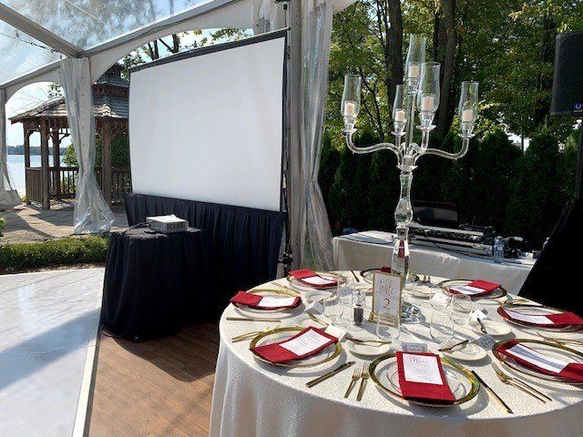 Outdoor event setup: tables with place settings, candelabra, projector screen, and lake view.