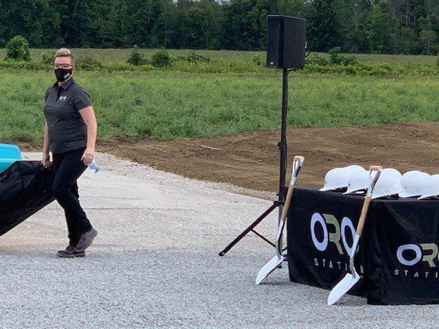 Woman in mask carrying a bag walks past a table with hard hats and shovels. Green field in the background.