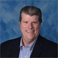 A man in a suit and striped shirt is smiling in front of a blue background.