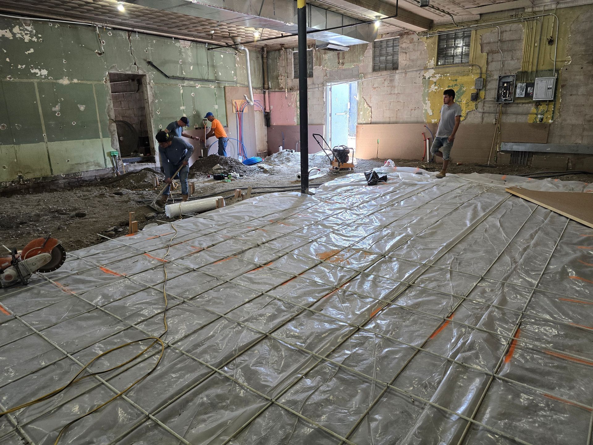 Workers laying plastic sheeting over a dirt floor in a worn indoor space during renovation