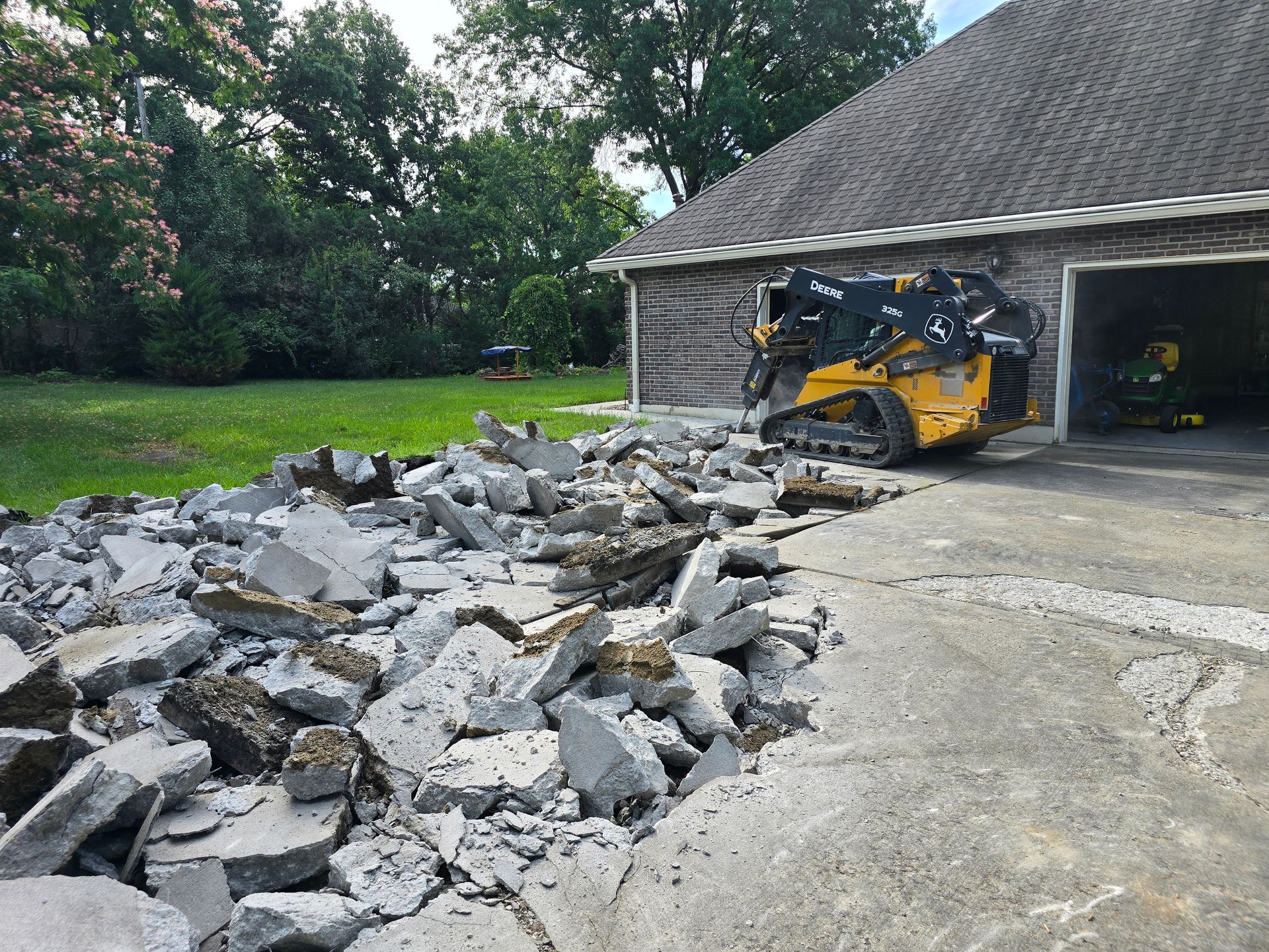 Pile of broken concrete and rocks beside a yellow skid steer in a driveway near a garage.