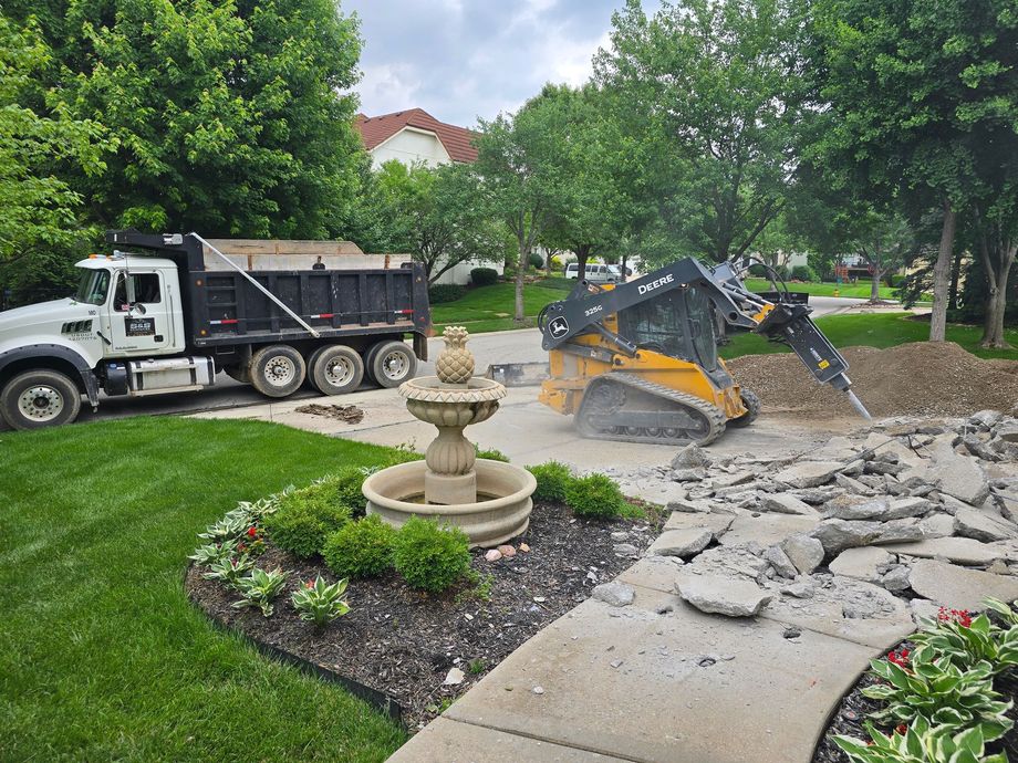 Construction equipment working on a concrete driveway beside a stone fountain and black dump truck