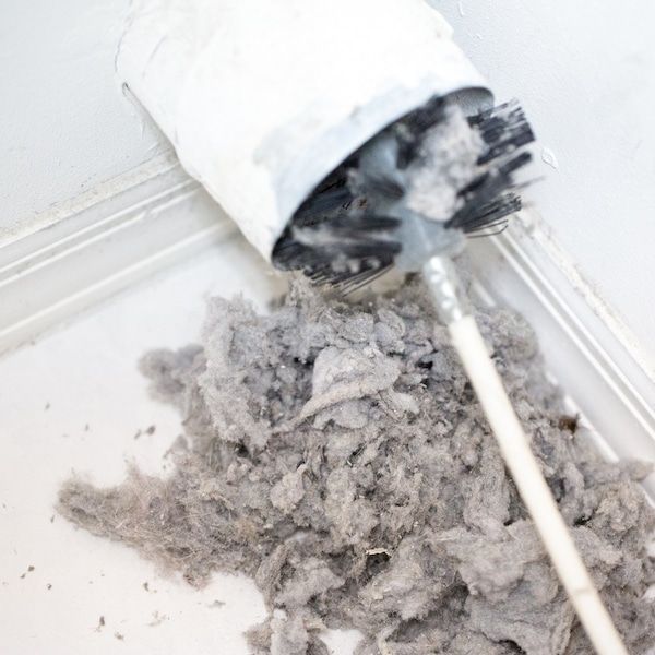 Hand holding a red hose with a green brush cleaning a dirty air vent in a white ceiling.