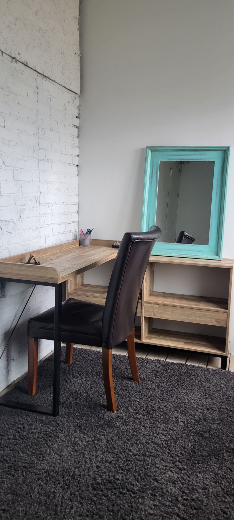 Small room with wooden desk, chair, turquoise-framed mirror, and shelves against a white brick wall.