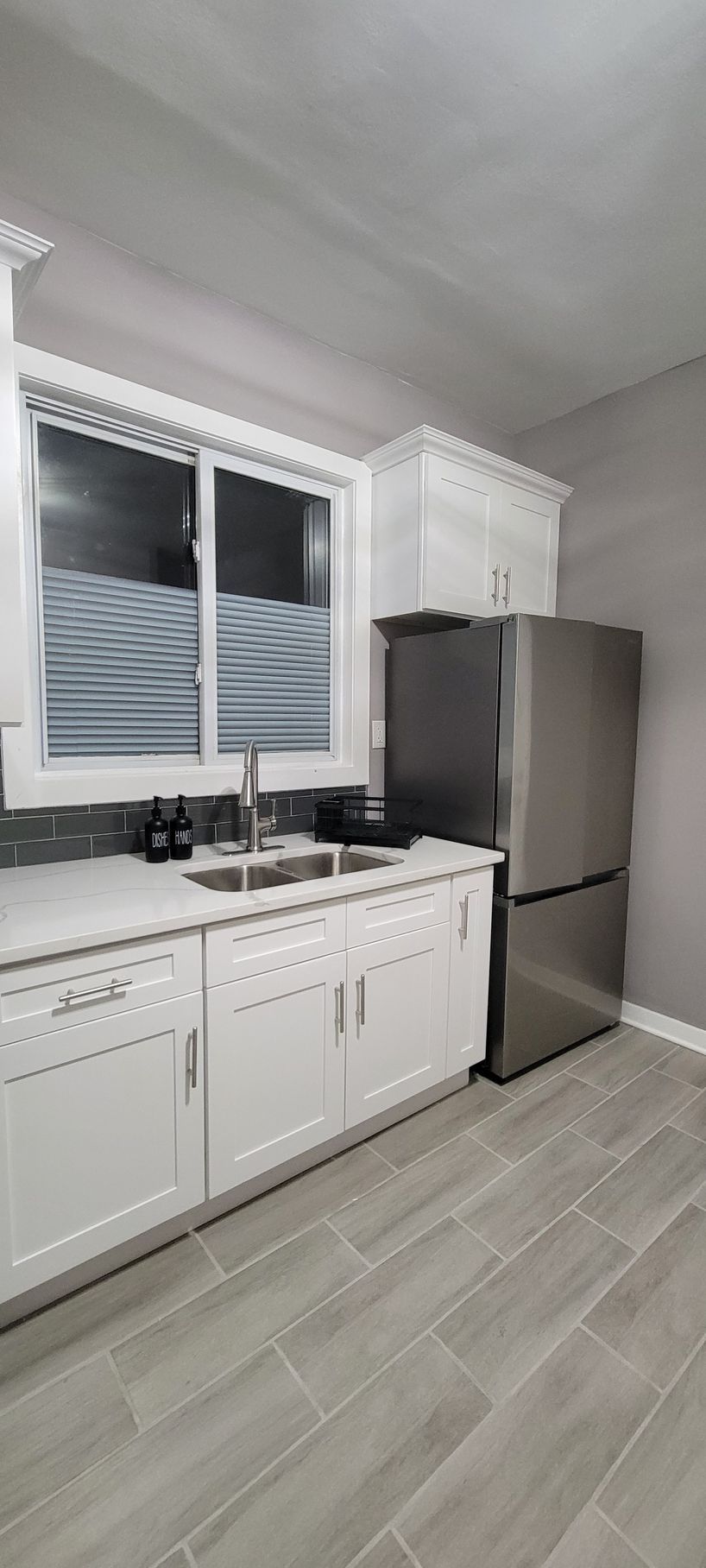 Modern white kitchen with stainless steel refrigerator, sink under window, and light wood-look tile floor