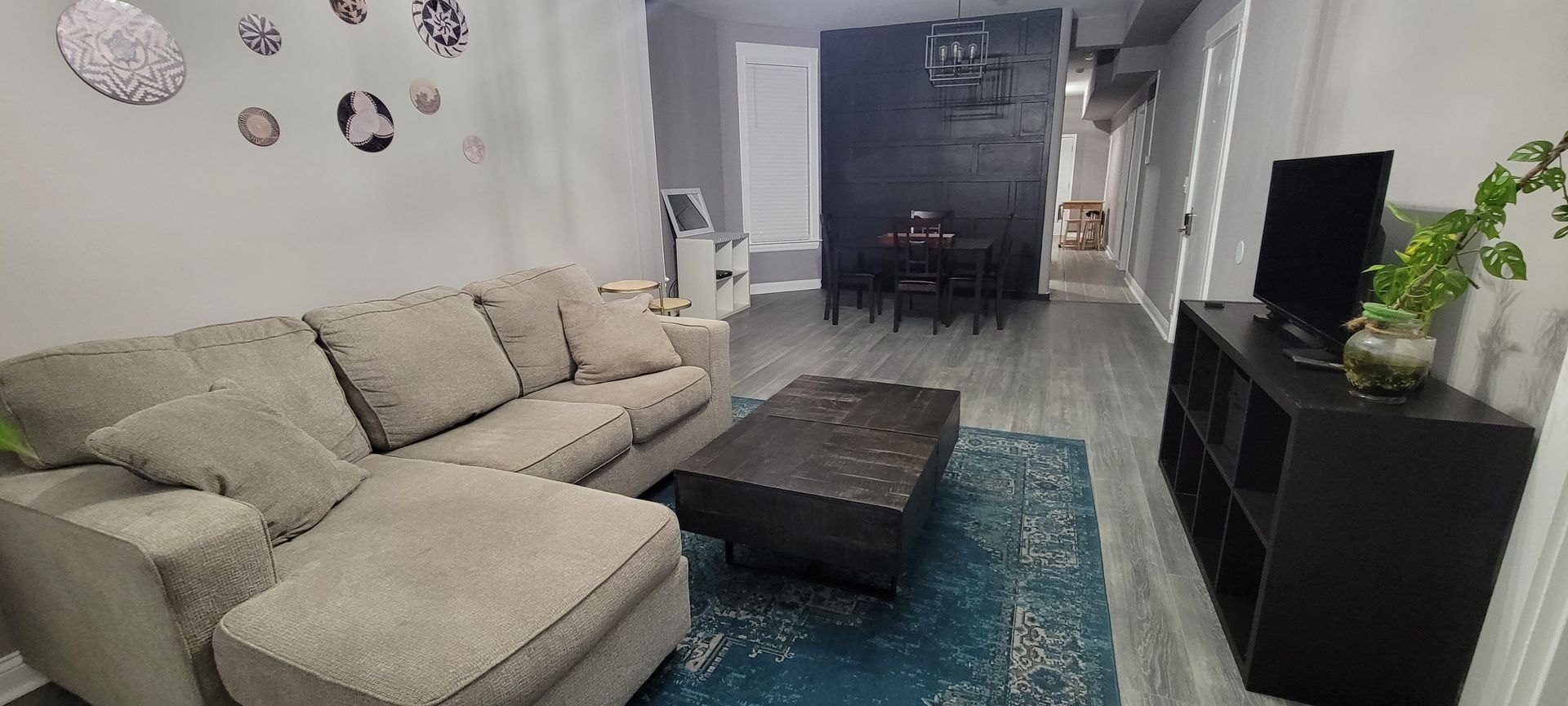 Modern living room with beige sectional sofa, dark coffee table, blue rug, and black TV console.