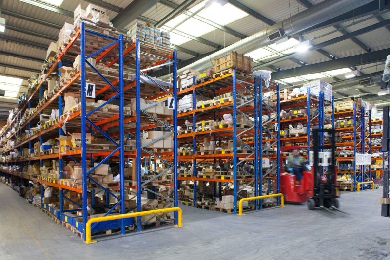Large warehouse interior with tall orange-and-blue shelving loaded with boxes and a forklift on the floor
