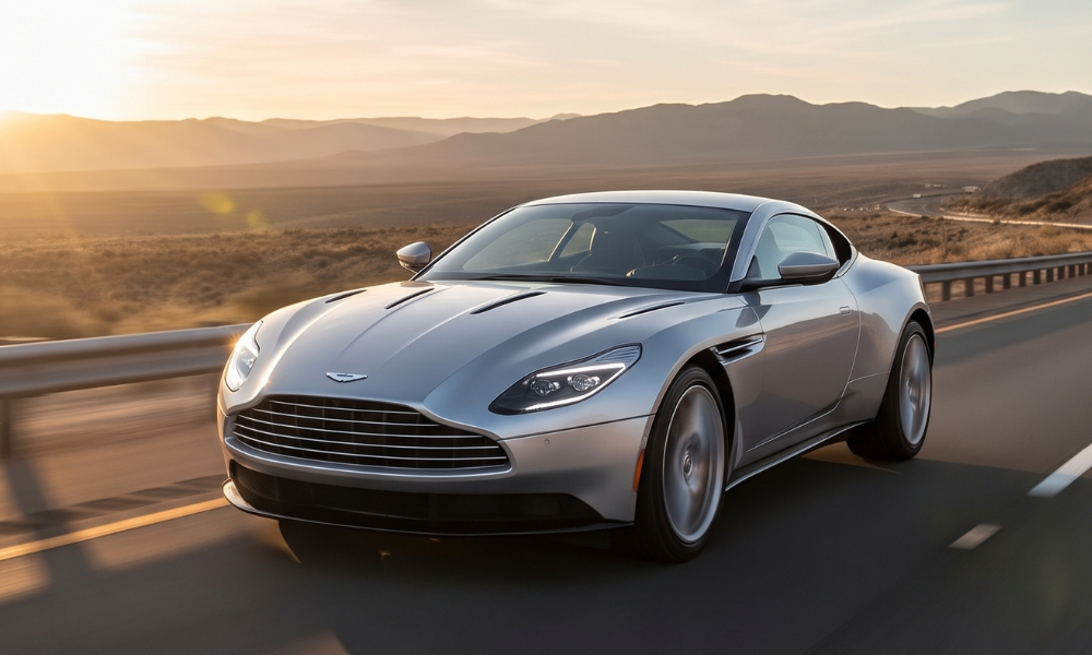 Silver Aston Martin grand touring sports coupe driving on a scenic desert highway at sunset, with warm sunlight illuminating the surrounding mountains