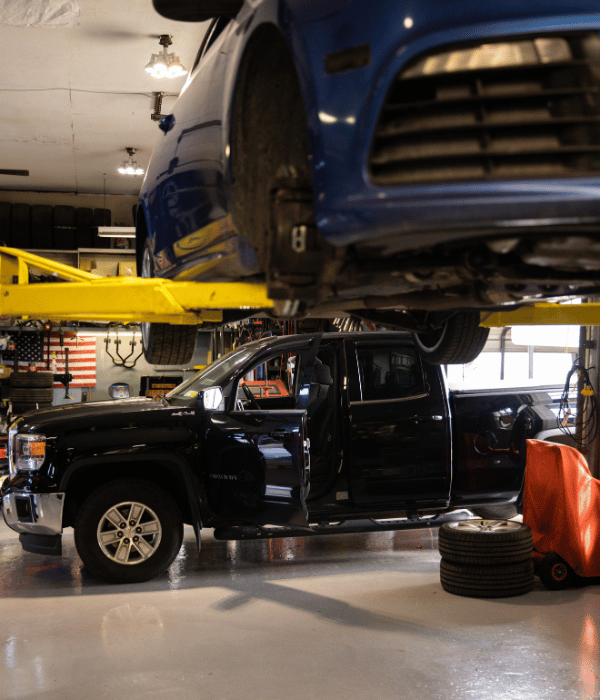 Pickup truck inside an automotive repair shop with another vehicle raised on a lift overhead.