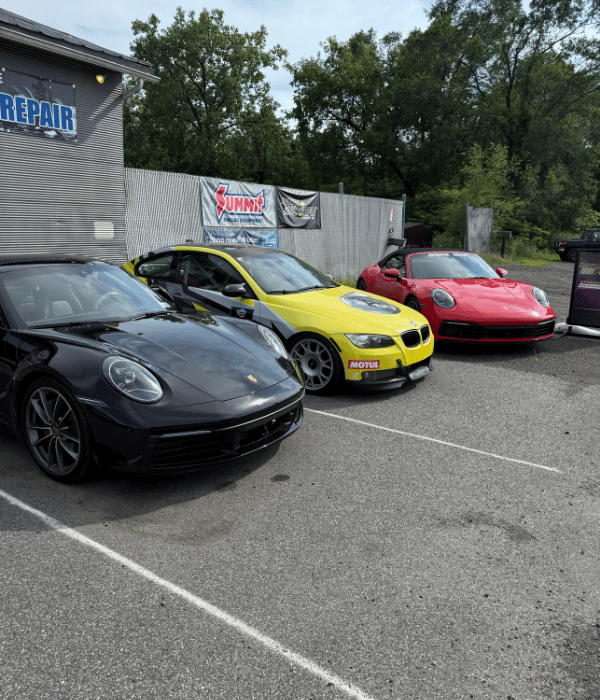 Black Porsche, yellow BMW race car, and red Porsche parked outside an automotive repair shop.